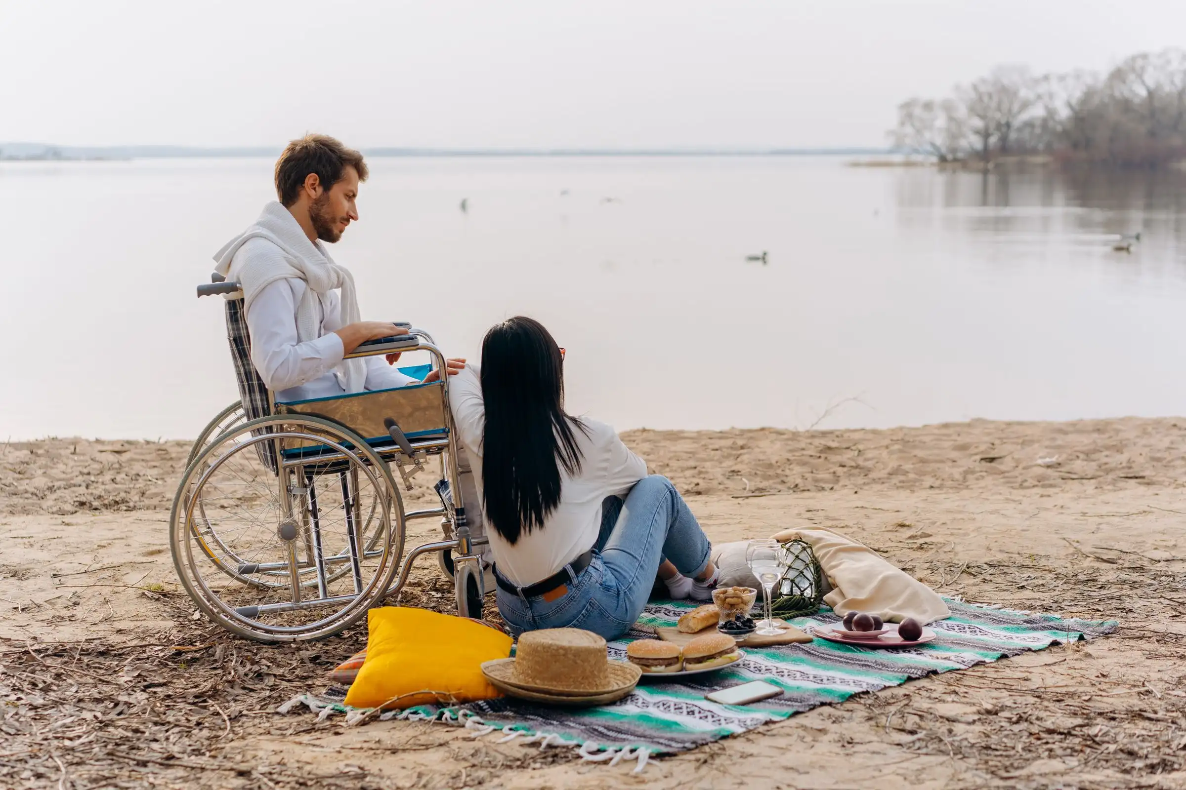 A man in a wheelchair and a friend having a picnic on a lakeside beach