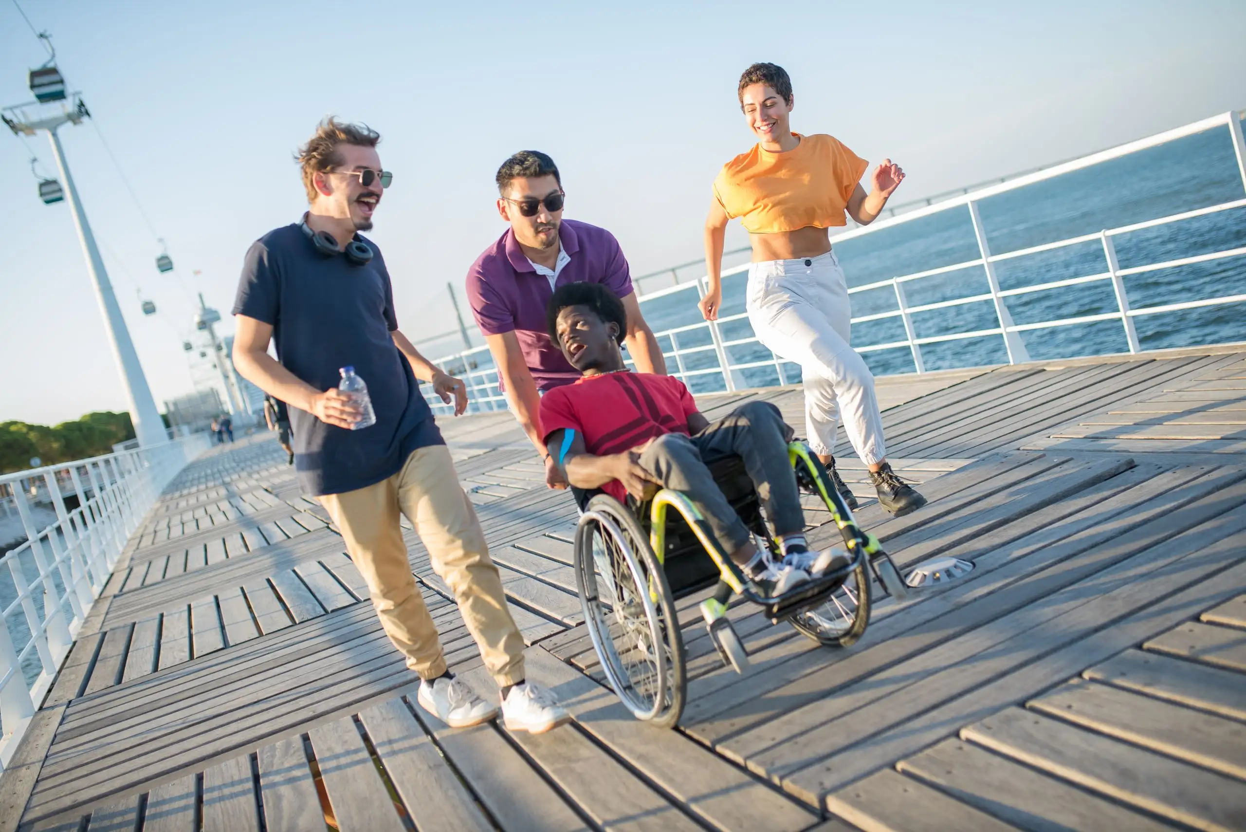 Four friends laughing together on a seaside boardwalk, one of them using a wheelchair