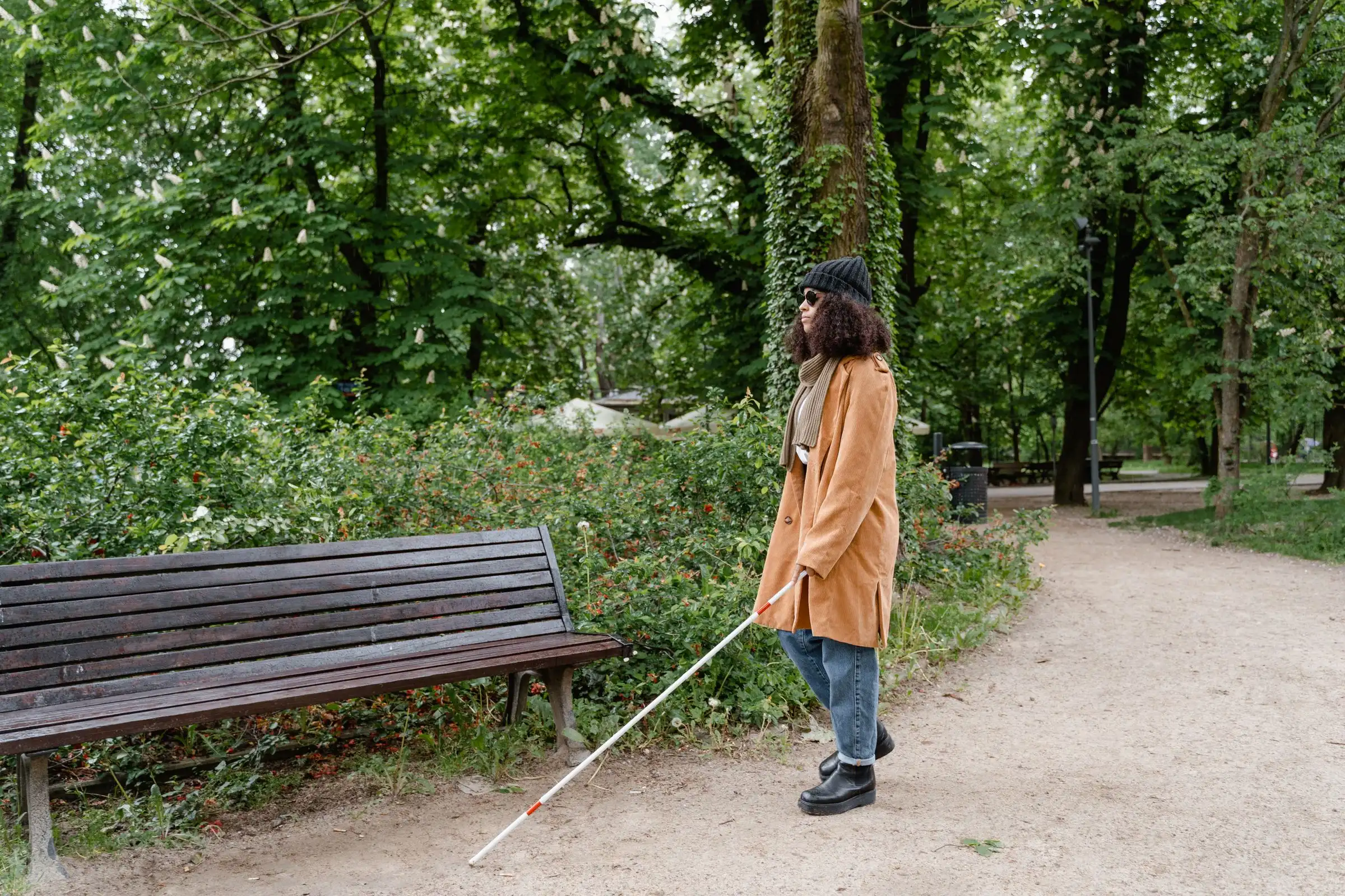 A woman with a white cane walking along a tree-lined park path